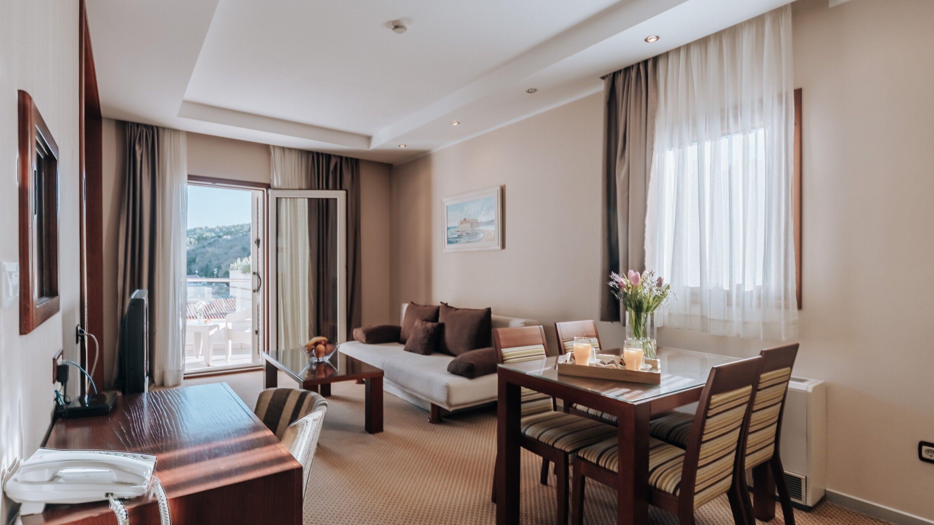 A hotel room with a dining area, a sofa, a TV, and a window with curtains.
