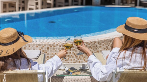 Dos personas brindando con copas de vino en una mesa junto a la piscina, con un libro y una taza.
