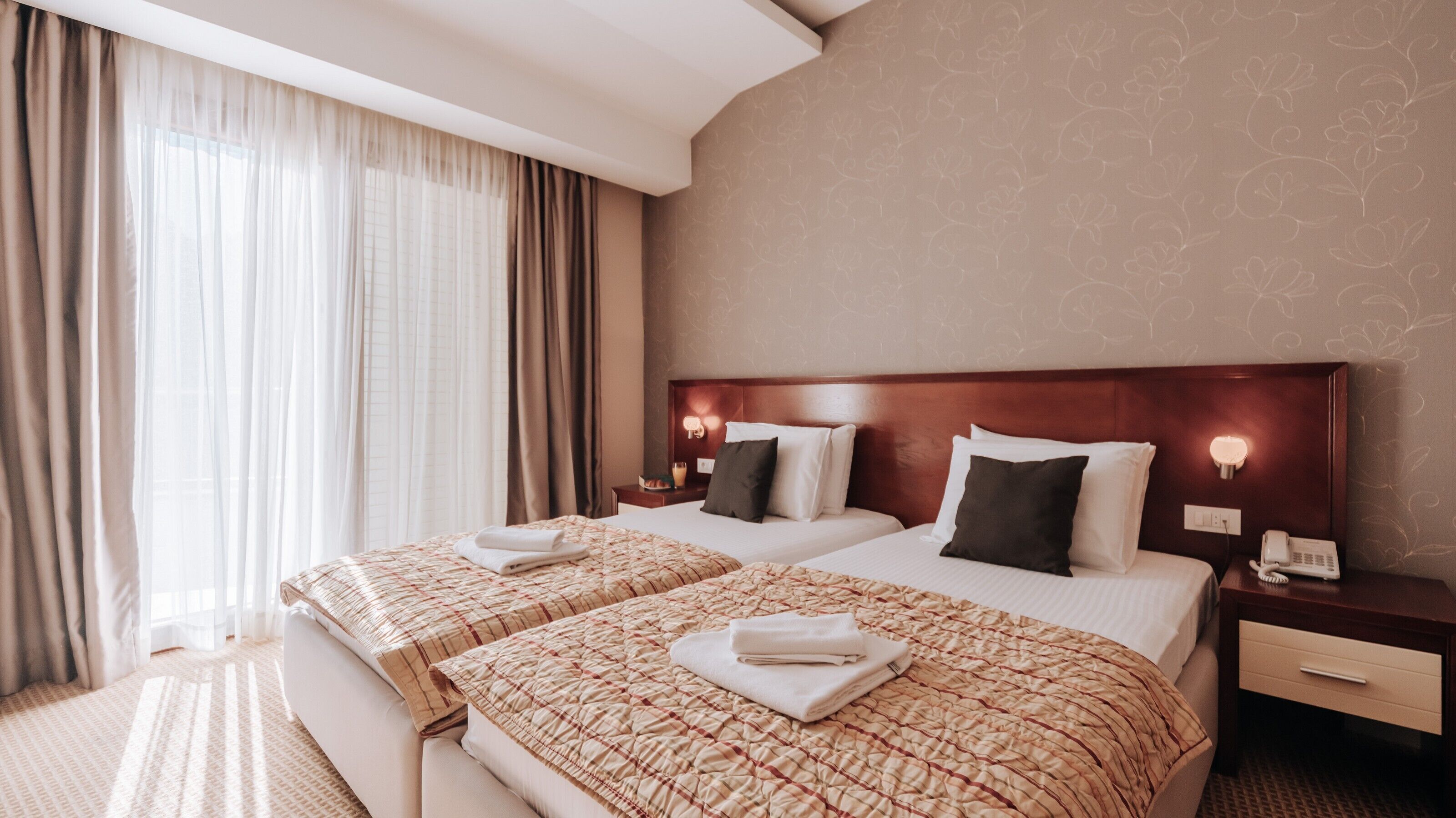A hotel room with two beds, each with a patterned bedspread and pillows, a wooden headboard, bedside lamps, and a telephone on the nightstand.