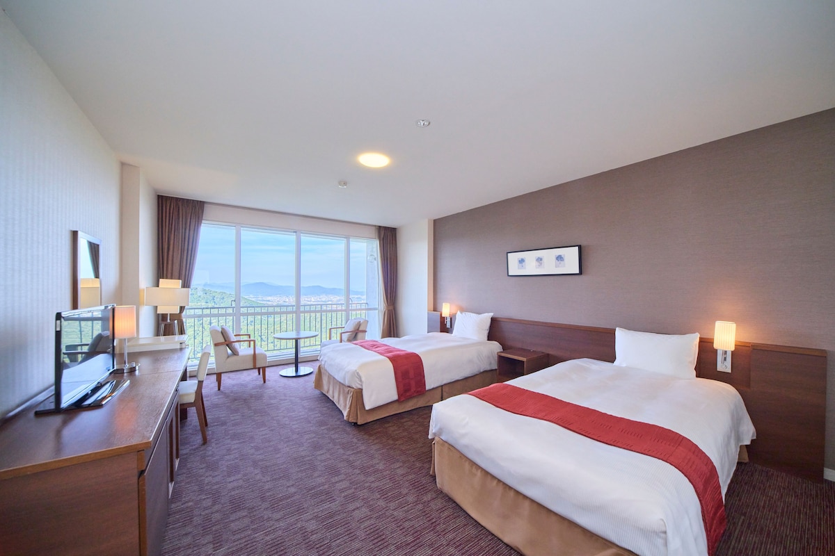 Habitación de hotel con dos camas, un escritorio, una silla y un amplio ventanal con vistas.