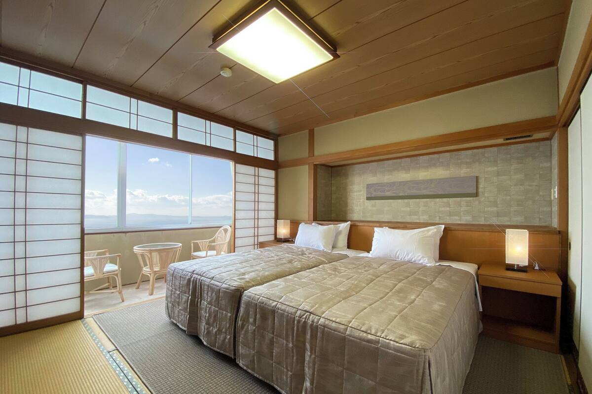 Quarto de hotel com duas camas, vista para o mar, piso de esteira de tatame e parede em estilo tradicional japonês.