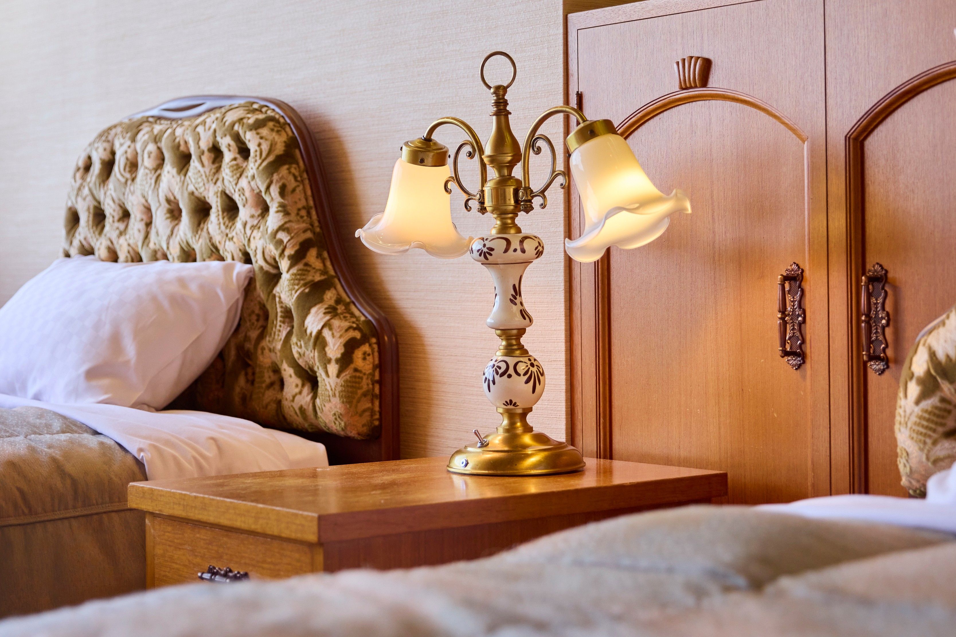 Una cama con cabecera capitonada, una mesita de noche con lámpara y un armario de madera con pomo.