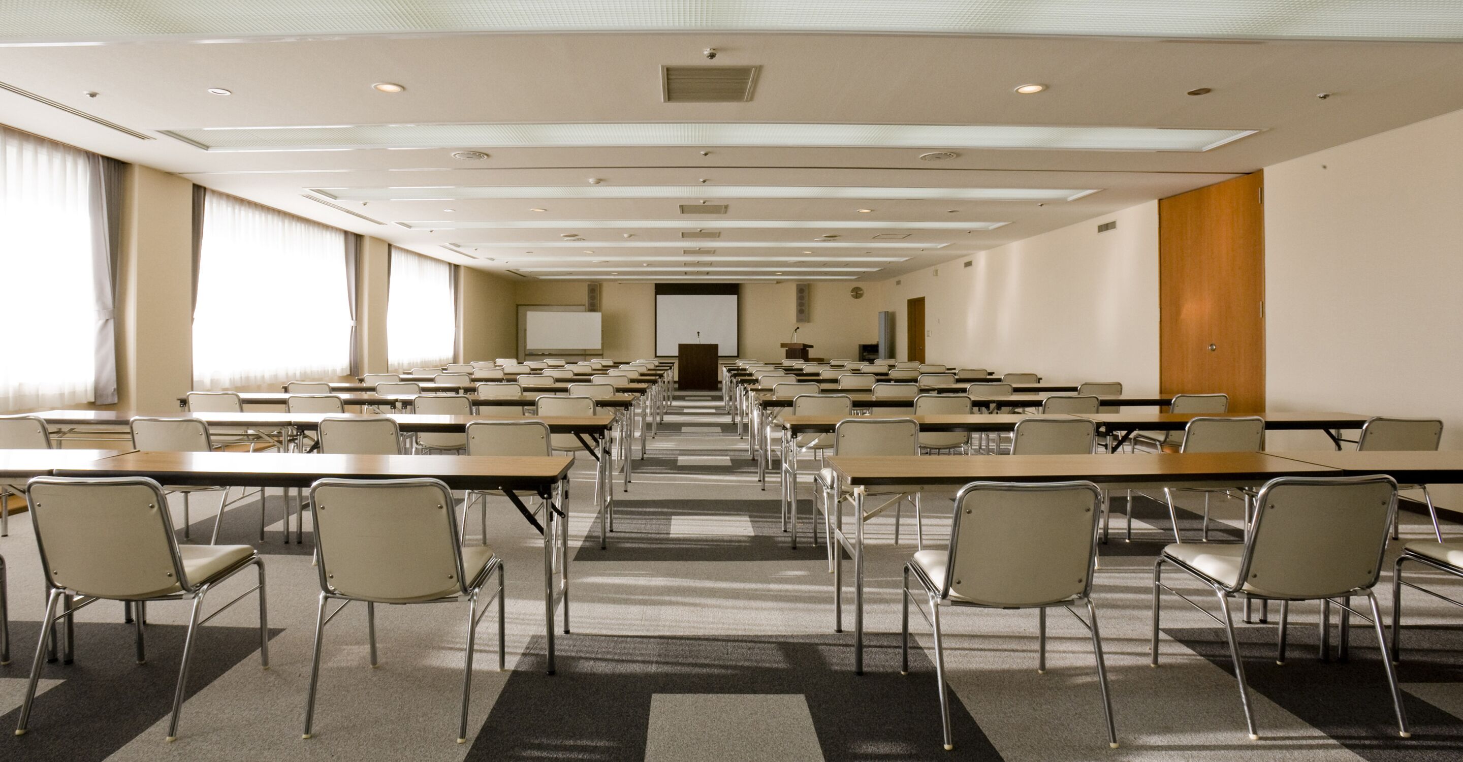 Une salle de conférence avec des rangées de tables et de chaises, un écran de projection et un pupitre.
