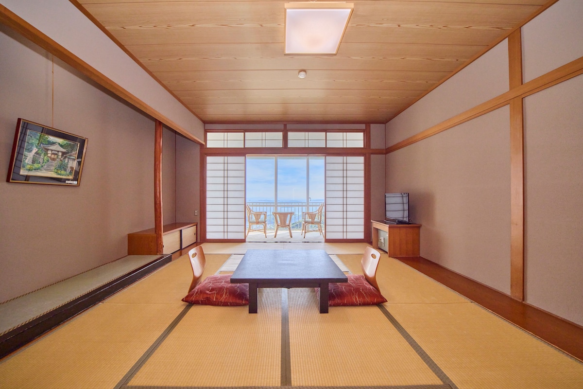 Habitación con tatami, una mesa baja, sillas, televisor y vistas a través de un celosía japonesa.