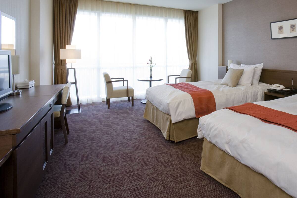 Quarto de hotel com duas camas, uma escrivaninha, uma cadeira, uma mesinha com um vaso e uma televisão.
