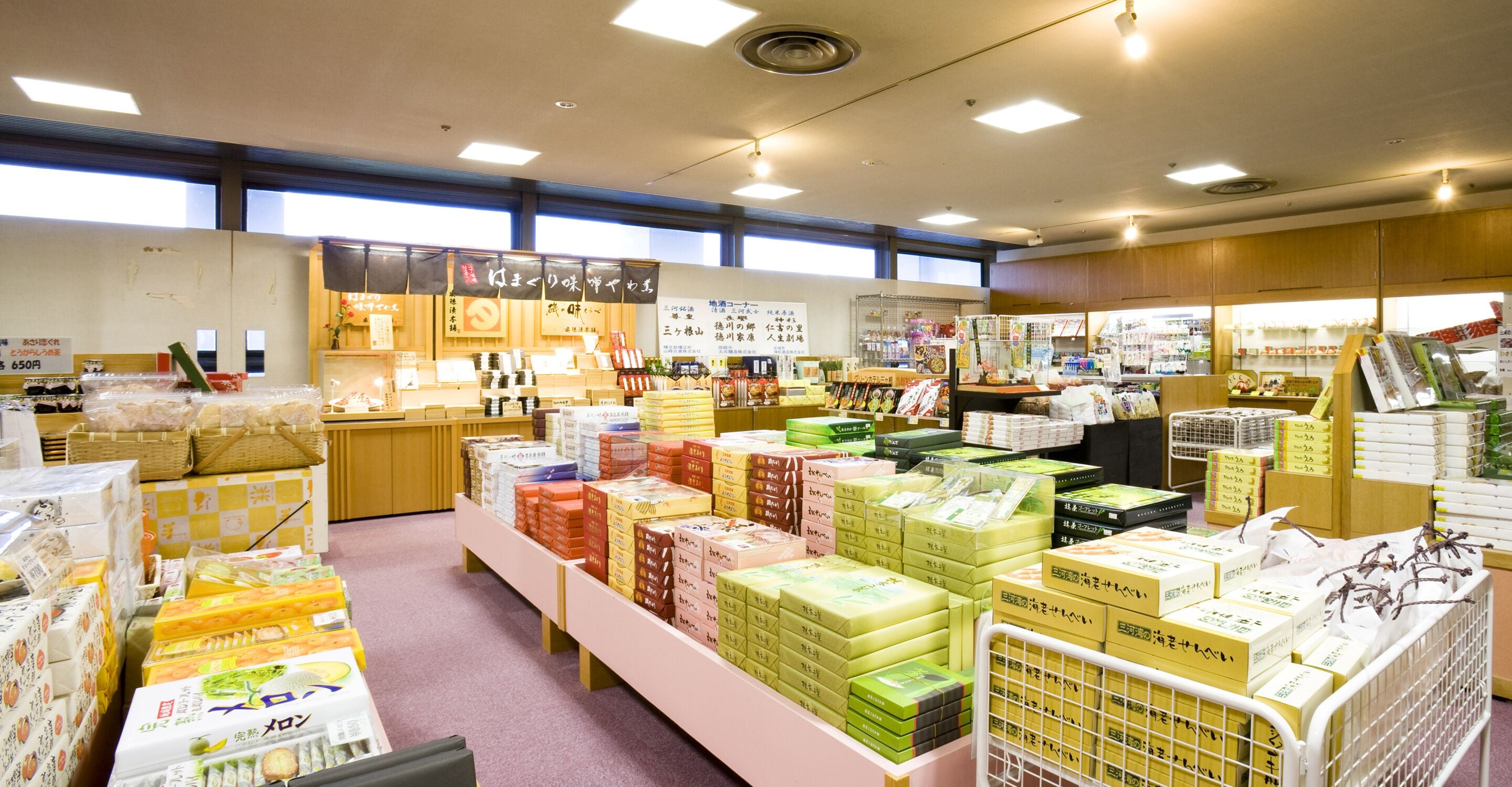 A store with various packaged goods on shelves and tables, wooden cabinets, and a windowed wall.