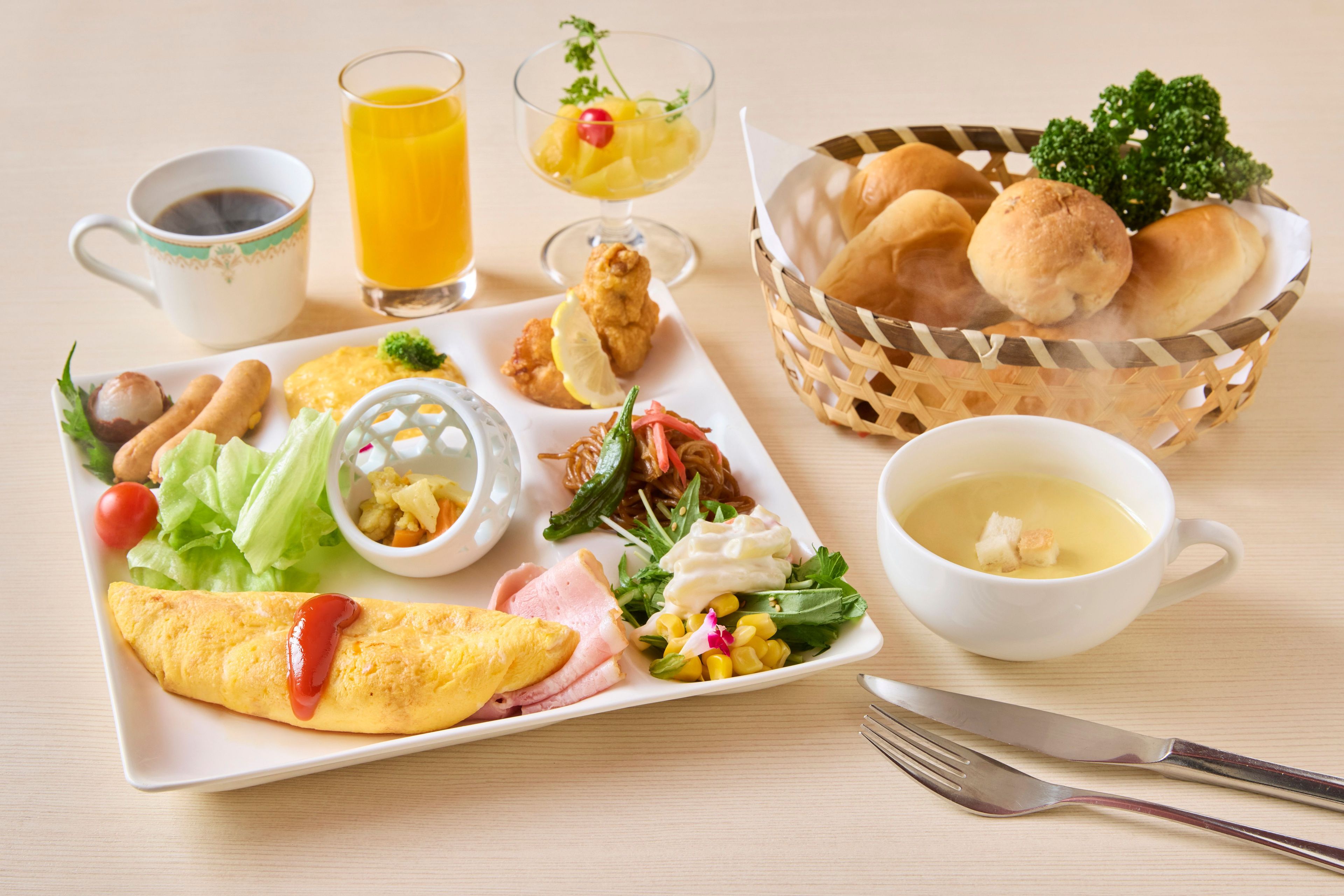 Une table mise pour le petit-déjeuner avec une assiette de nourriture, un bol de soupe, un panier de pain, une tasse de café et un verre de jus d’orange.