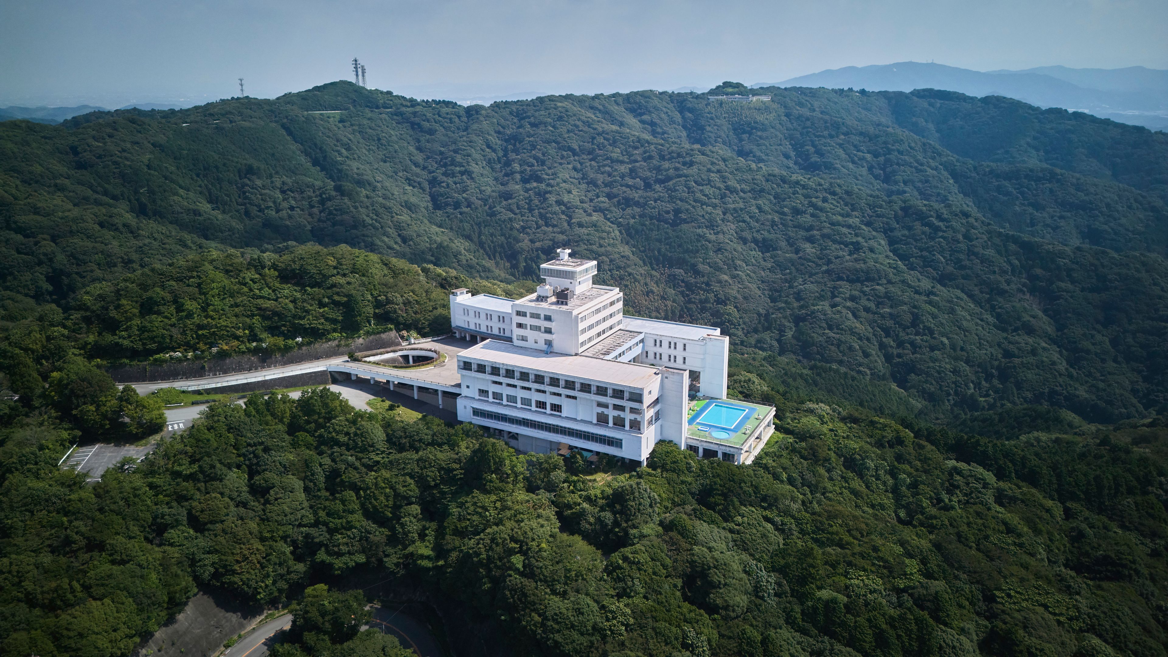A large, multi-story building with a flat roof and a swimming pool, surrounded by dense forest and hills.