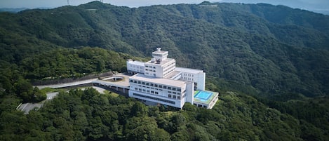 Un edificio grande de varios pisos con techo plano y piscina, rodeado de un bosque denso y colinas.