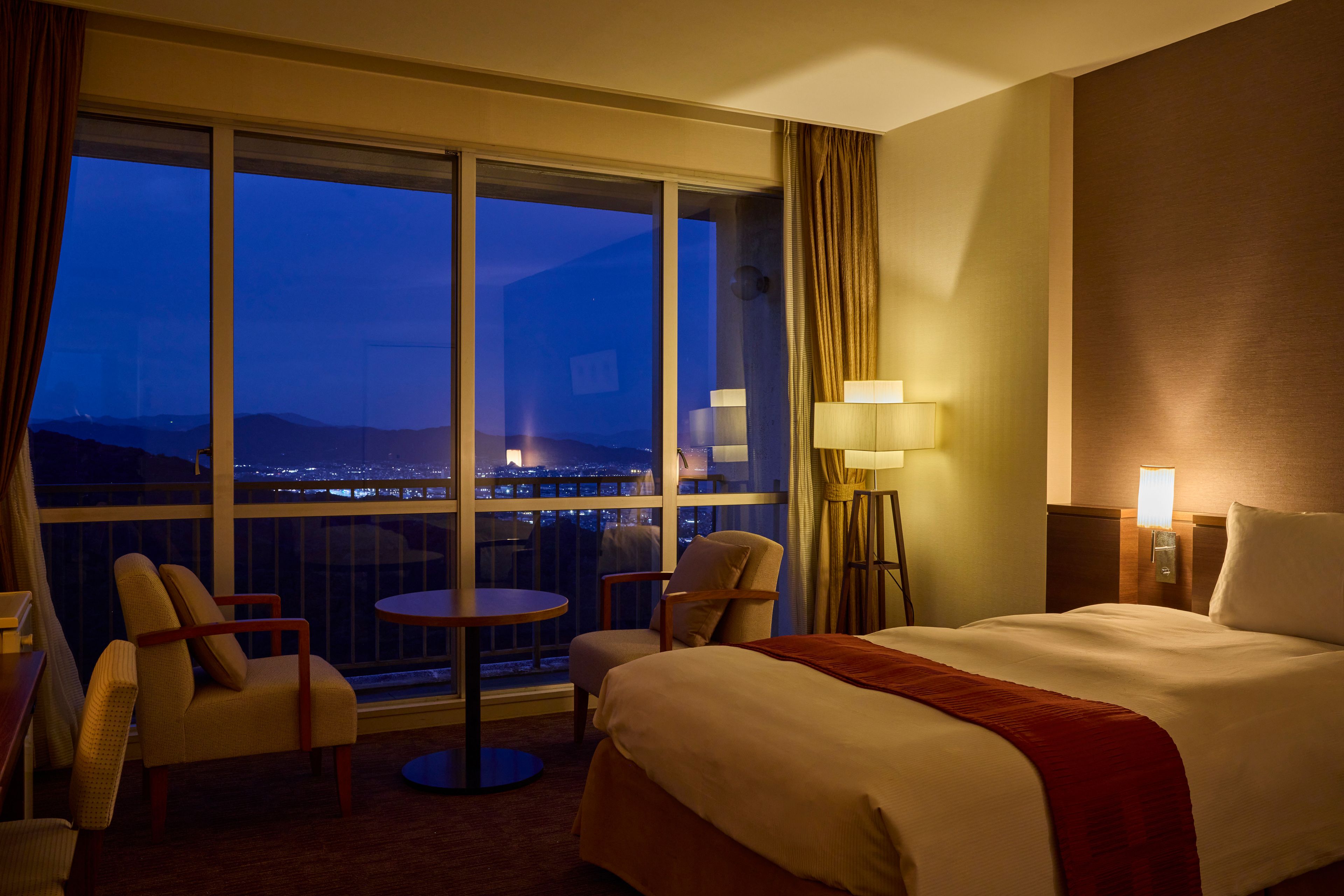 Ein Hotelzimmer mit großem Fenster, einem Bett, einem Tisch, zwei Stühlen und einer Lampe.