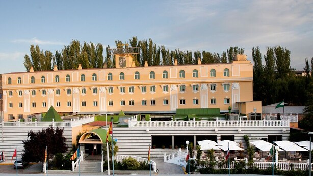 Un grand hôtel à plusieurs étages, doté d’une façade symétrique, d’une entrée centrale avec des marches et d’un espace extérieur couvert agrémenté de parasols.