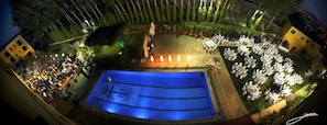 A pool area with tables and chairs arranged for an event, surrounded by greenery and buildings.