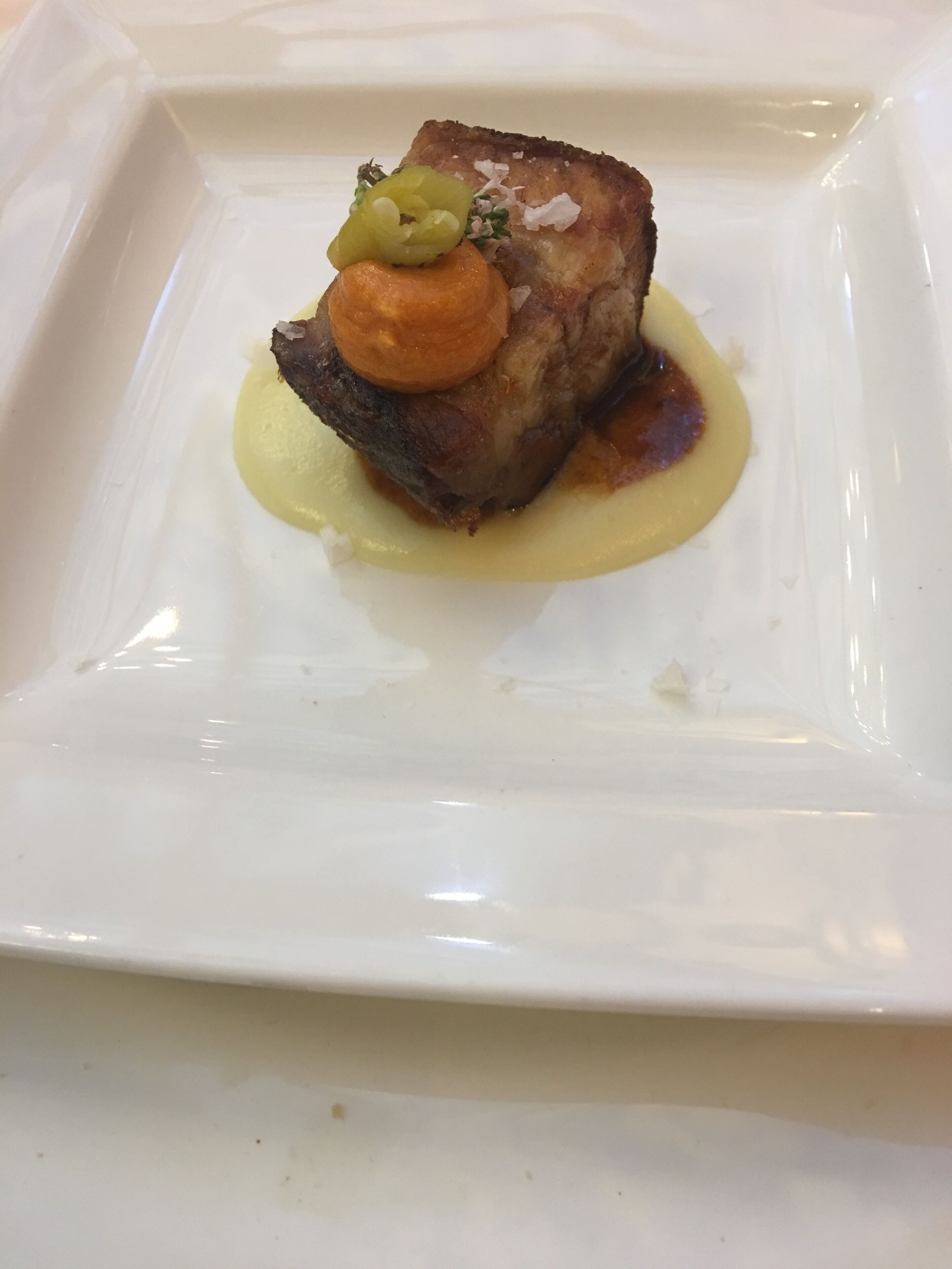 A square white plate with a single piece of meat, a garnish, and a sauce.