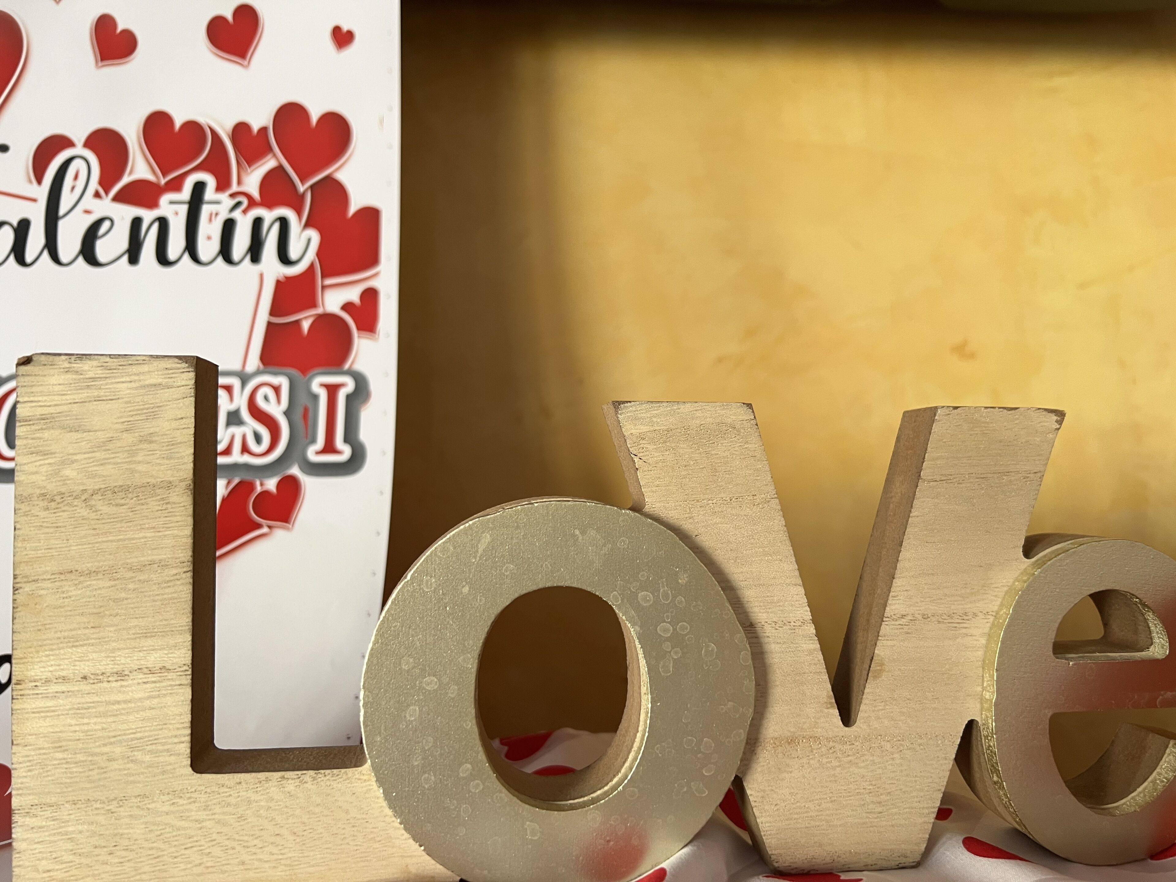 Des lettres en bois formant le mot « AMOUR », avec une finition dorée, placées devant un panneau décoré sur le thème de la Saint-Valentin, orné de cœurs et du mot « Valentin ».