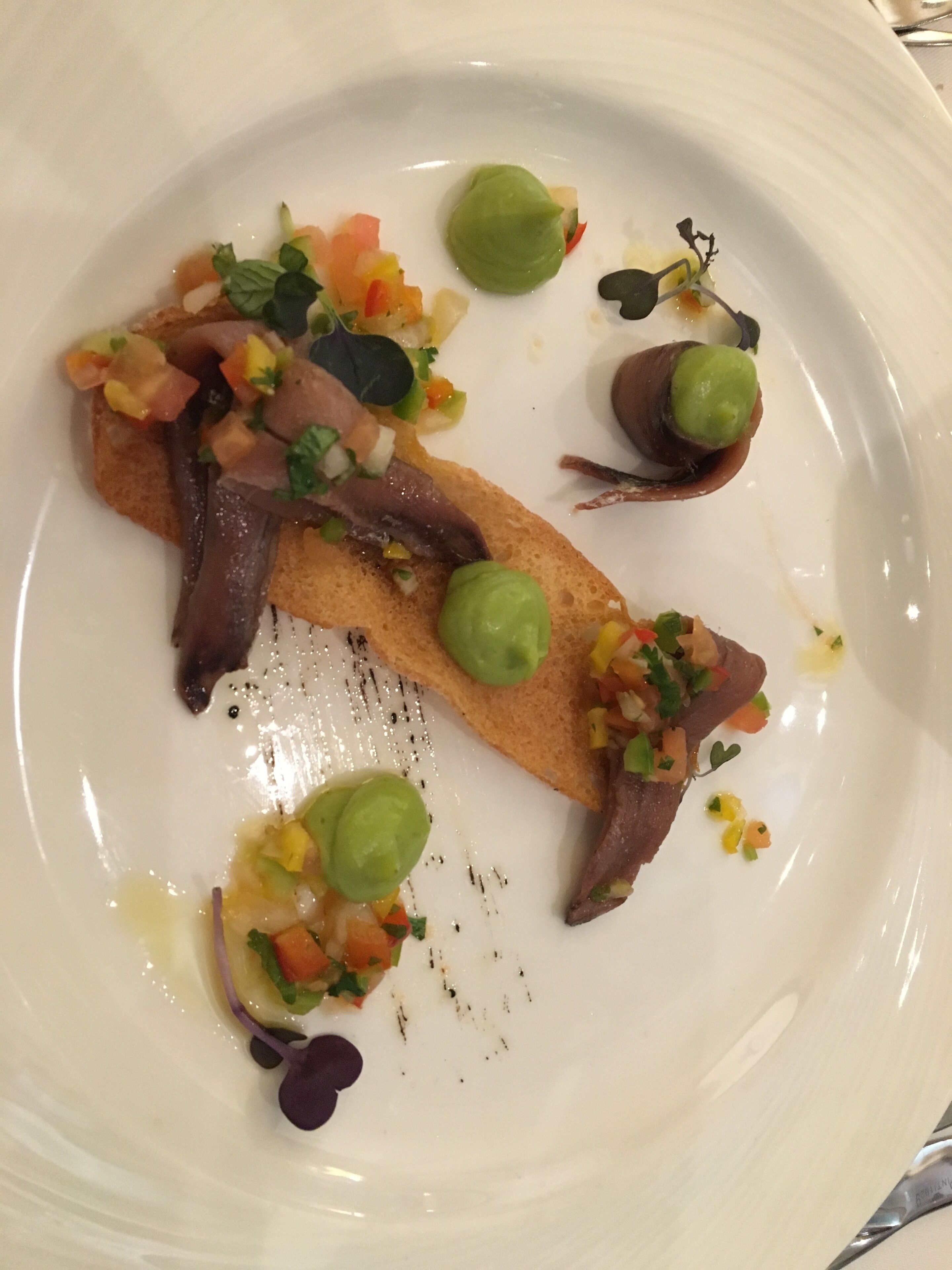 A white plate with a piece of bread topped with a dark sauce, green leaves, and a colorful vegetable topping.
