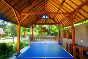 Una mesa de ping pong bajo un pabellón de madera con techo de paja, rodeada de abundante follaje.