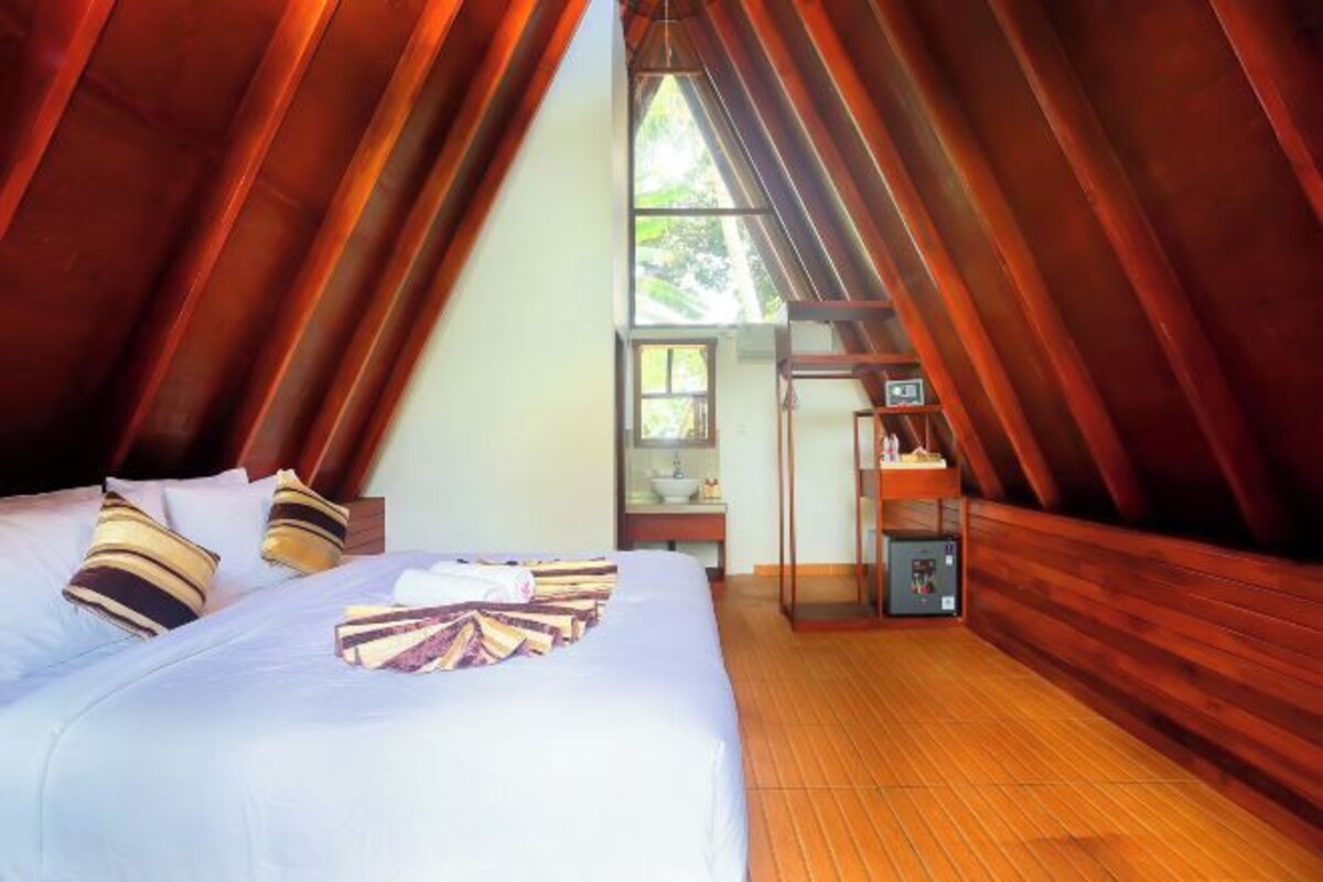 Una habitación acogedora de forma triangular, con techo y paredes de madera, una cama con ropa blanca y una pequeña mesa con una lámpara.