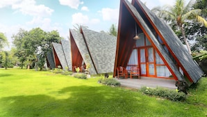 A row of modern, triangular-roofed buildings with a well-maintained lawn and palm trees in the background. - Dewantara Resort Bali (Gerokgak)
