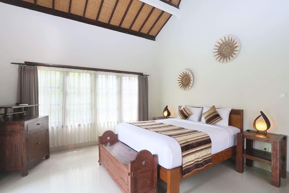 A bedroom with a wooden bed, a nightstand with a lamp, a dresser, and a large window with curtains.