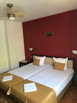 Een tweepersoonsbed met witte lakens, twee kussens en een bruin sprei. Er is een plafondventilator, twee bedlampjes en een klein schilderijtje aan de muur.