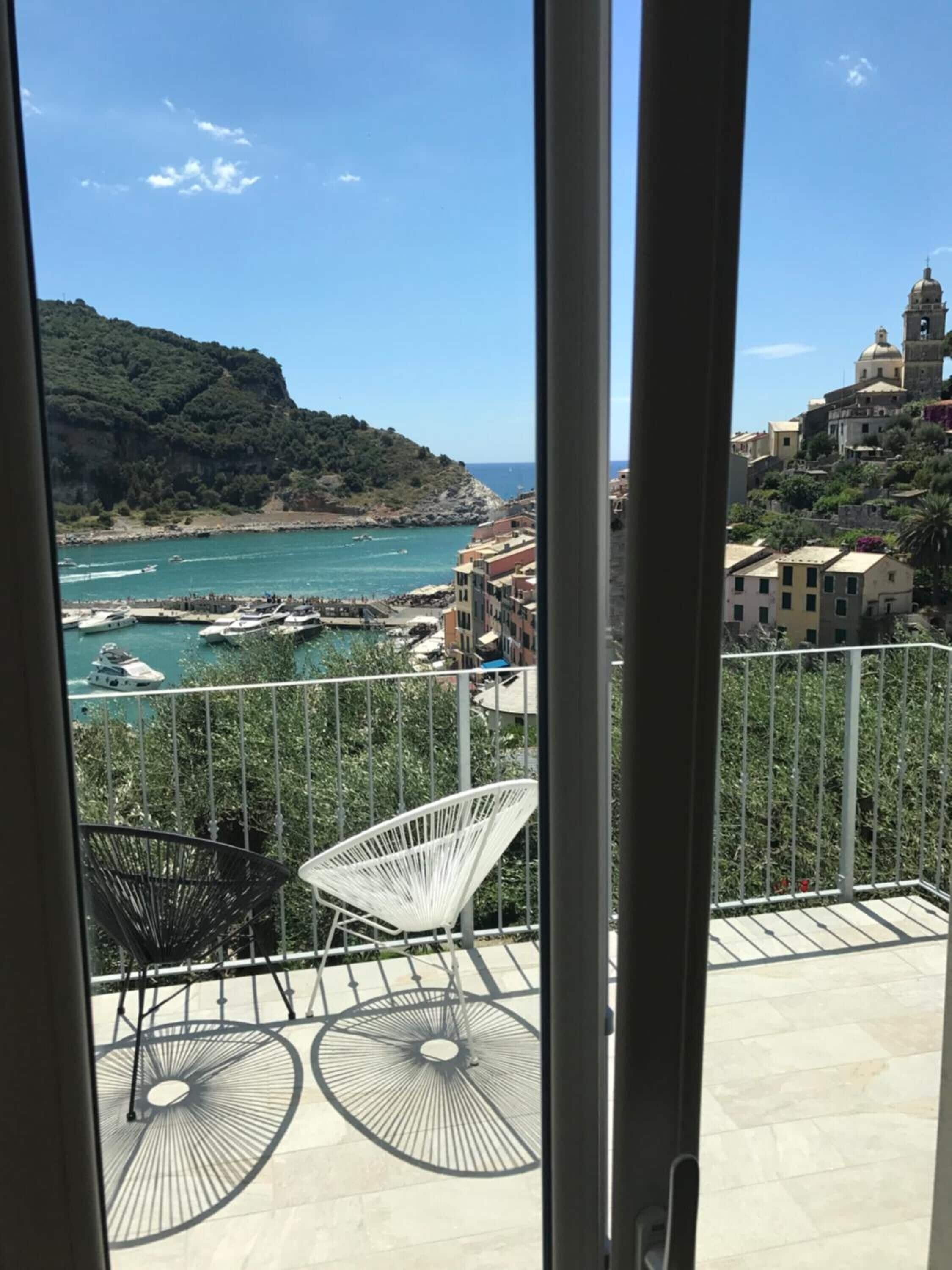 Un balcone con due sedie e un tavolo, affacciato su una località costiera con barche e una collina.