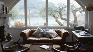 A cozy room with a large arched window, a leather sofa with cushions, a small table with a book, and a lamp on a side table. - Relais Santa Caterina (Portovenere)