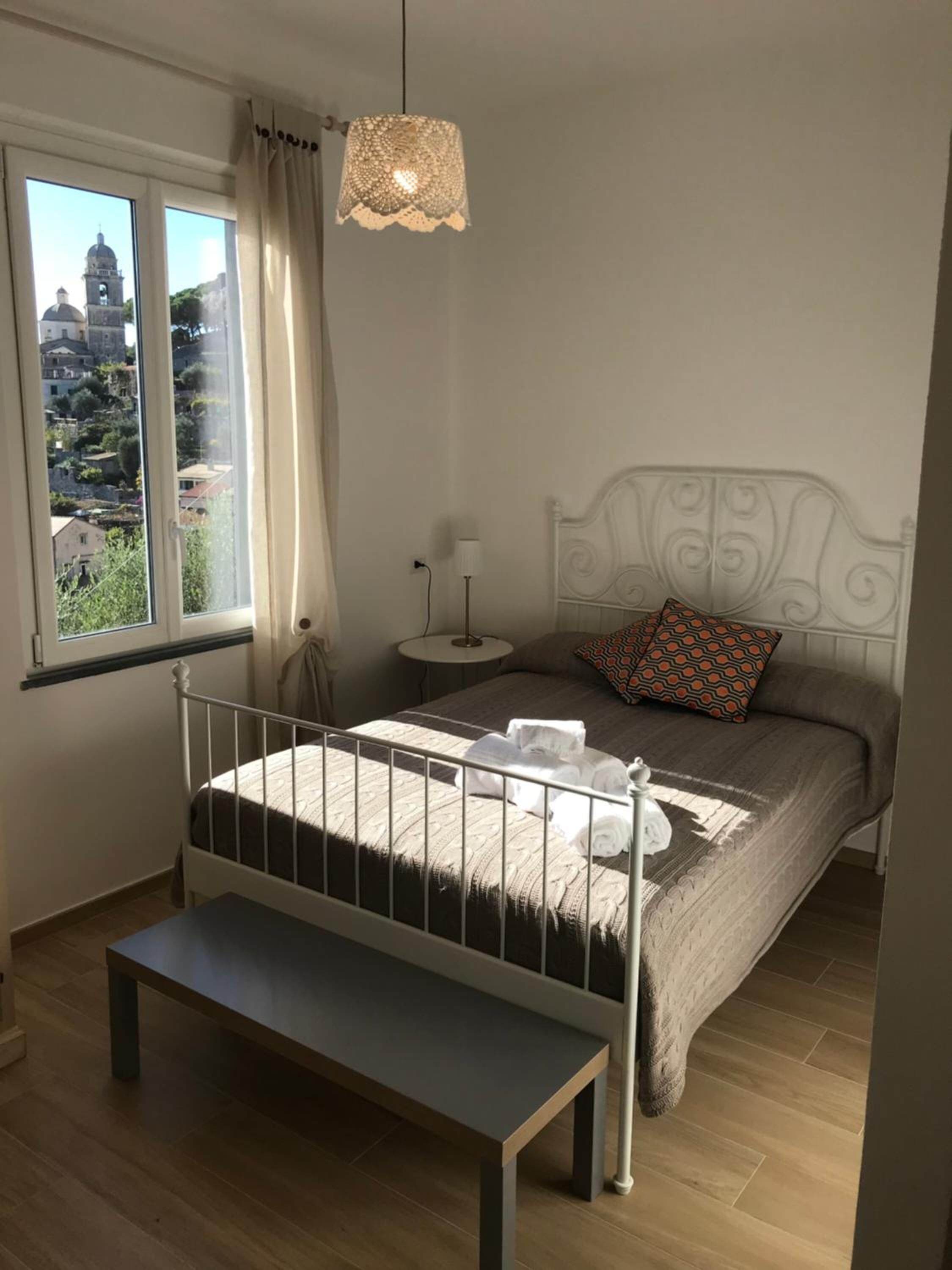 Ein Schlafzimmer mit Bett, Nachttisch, Lampe, Bank und Blick auf ein Gebäude durch das Fenster.