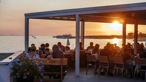 Un espace extérieur aménagé avec des tables et des chaises, offrant une vue sur un plan d’eau et un coucher de soleil en arrière-plan.