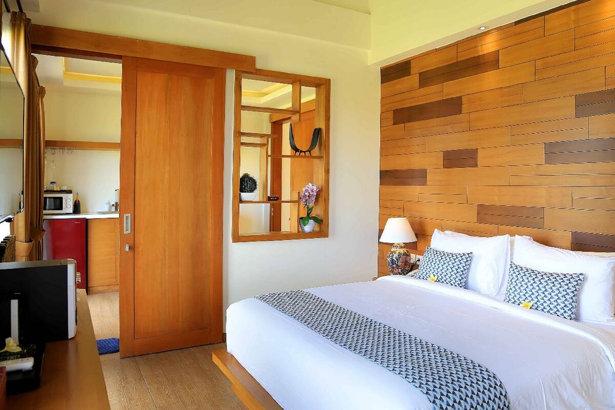 A hotel room with a wooden headboard, a large bed with patterned bedding, a bedside table with a lamp, and a view into the kitchen area.