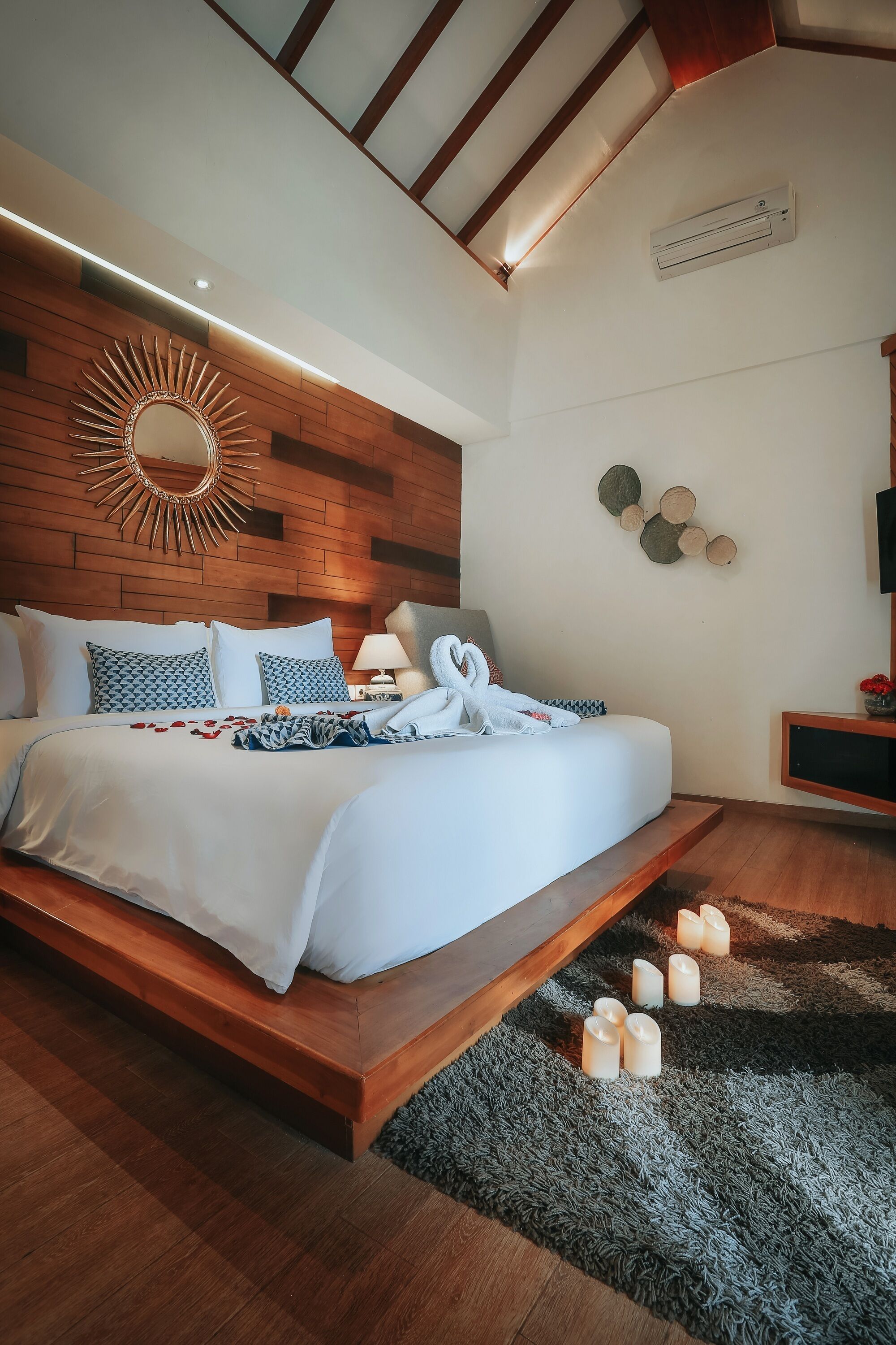 A modern bedroom with a wooden headboard, a large mirror, a bed with white linens, and a small bedside table with a lamp.