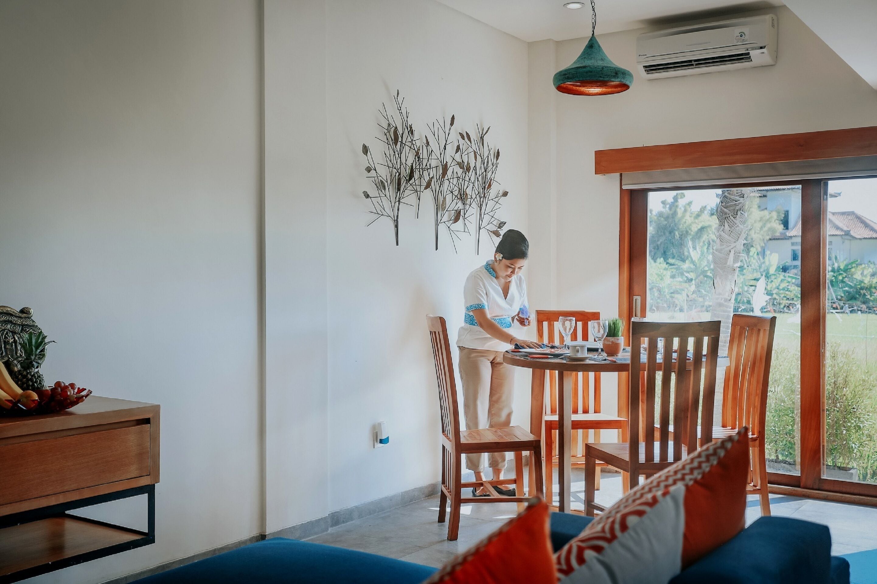 A modern dining area with a wooden table and chairs, a decorative wall piece, and a view of greenery outside.