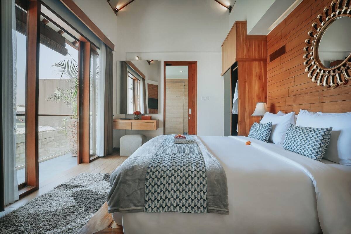A modern hotel room with a large bed, a wooden headboard, a mirror, and a view through large sliding doors.