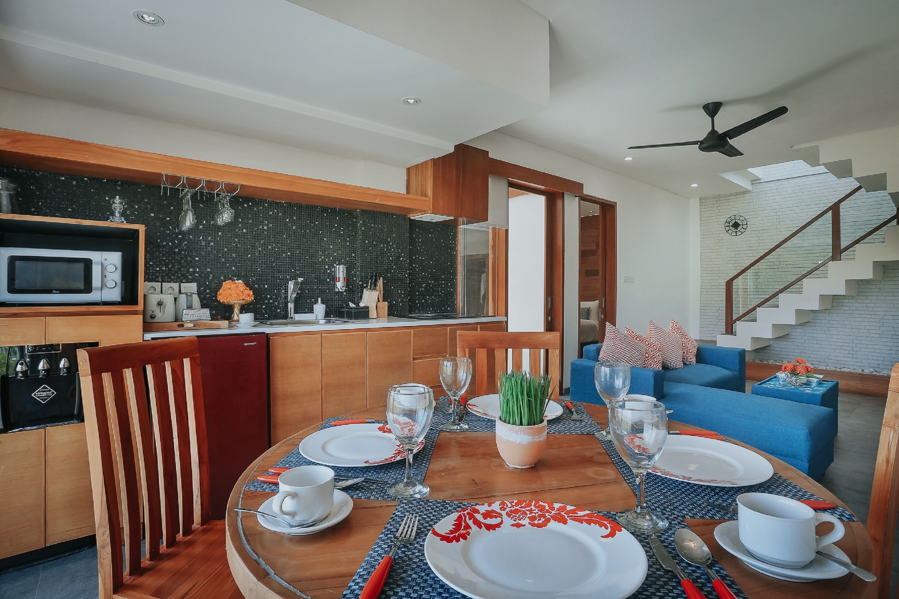 A modern dining area with a wooden table set for four, a kitchen with a microwave and sink, and a staircase leading to an upper level.