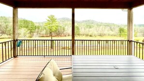 Un espacio al aire libre techado, con techo de madera, una mesa blanca y un cojín beige. Ofrece vistas a un paisaje verde con árboles y una valla.