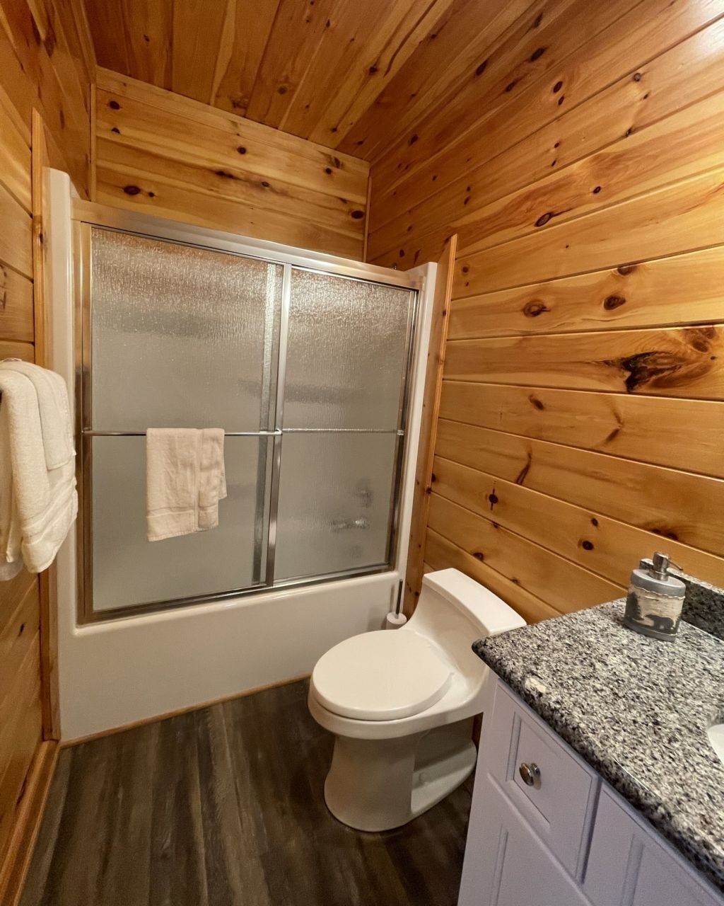 A compact bathroom with wooden walls, a glass shower enclosure, a white toilet, and a granite countertop with a sink.