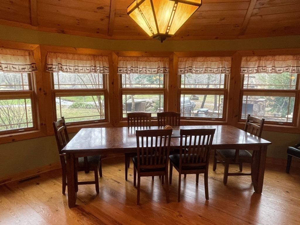 A dining room with a large wooden table and chairs, large windows with curtains, and a wooden ceiling.