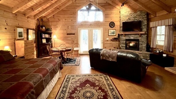Ein Holzblockhaus-Innenraum mit Holzbalkendecke, Kamin, Essbereich und einem Schlafzimmer mit großem Bett.