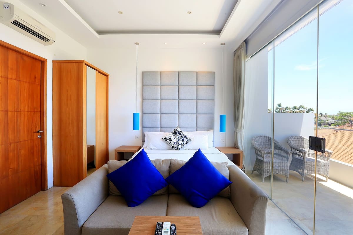 Une chambre d’hôtel moderne avec un grand lit, un canapé avec des coussins bleus, un balcon avec des fauteuils en osier et une vue sur le paysage urbain.