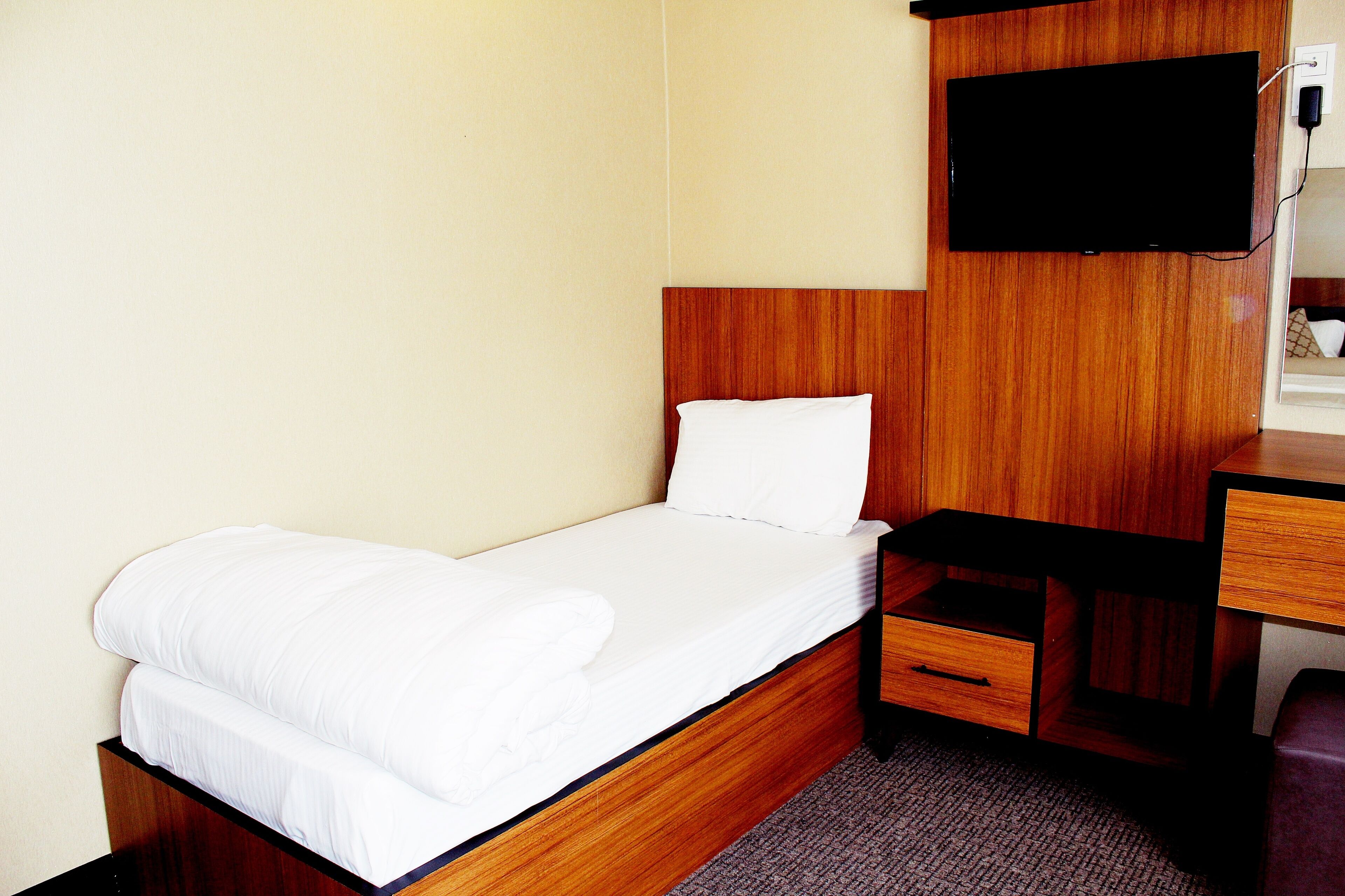 A single bed with a wooden headboard, a flat-screen TV mounted above it, and a desk with a chair to the right.