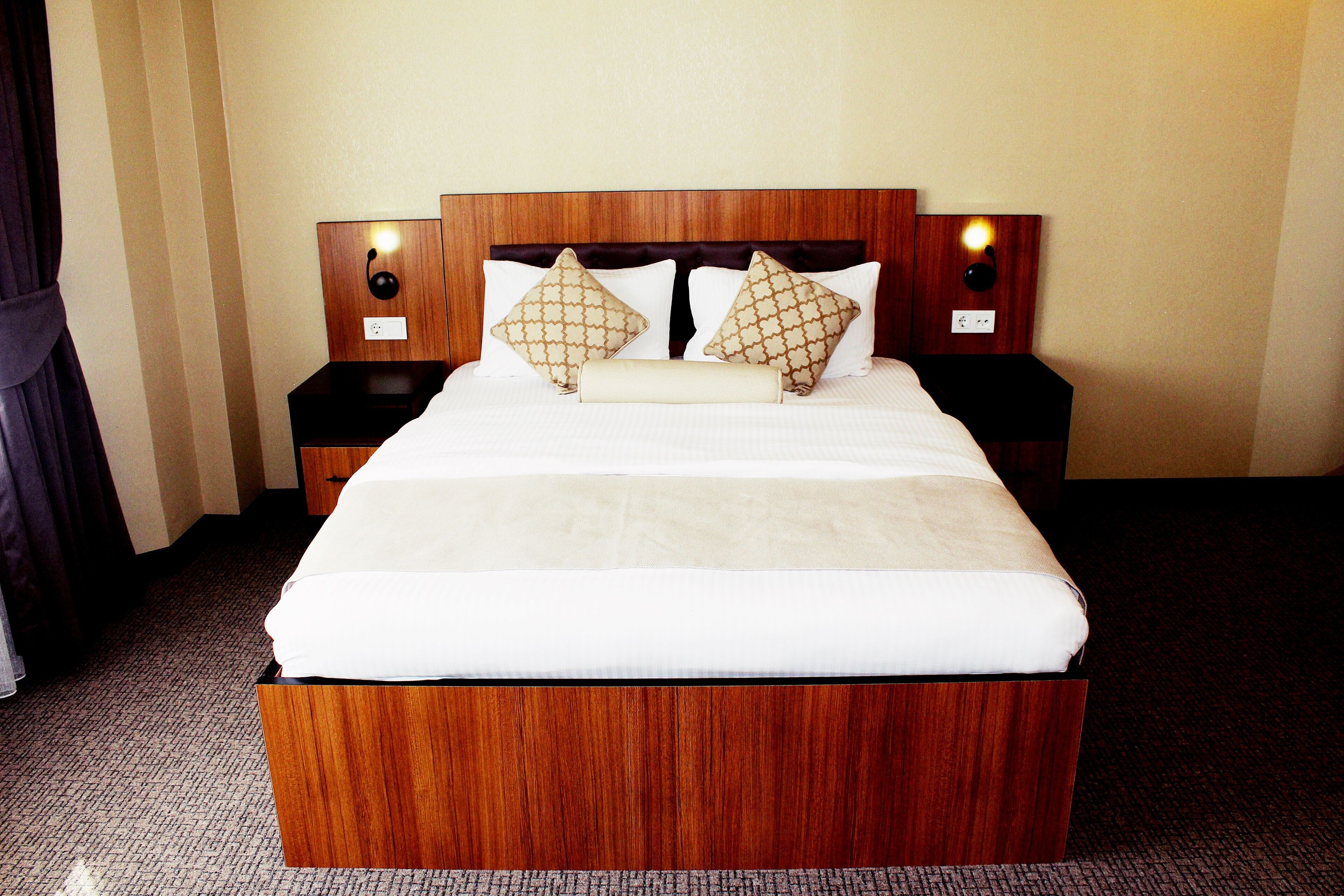 Una habitación de hotel con una cama de madera grande, dos mesitas de noche con lámparas y una cortina a la izquierda.