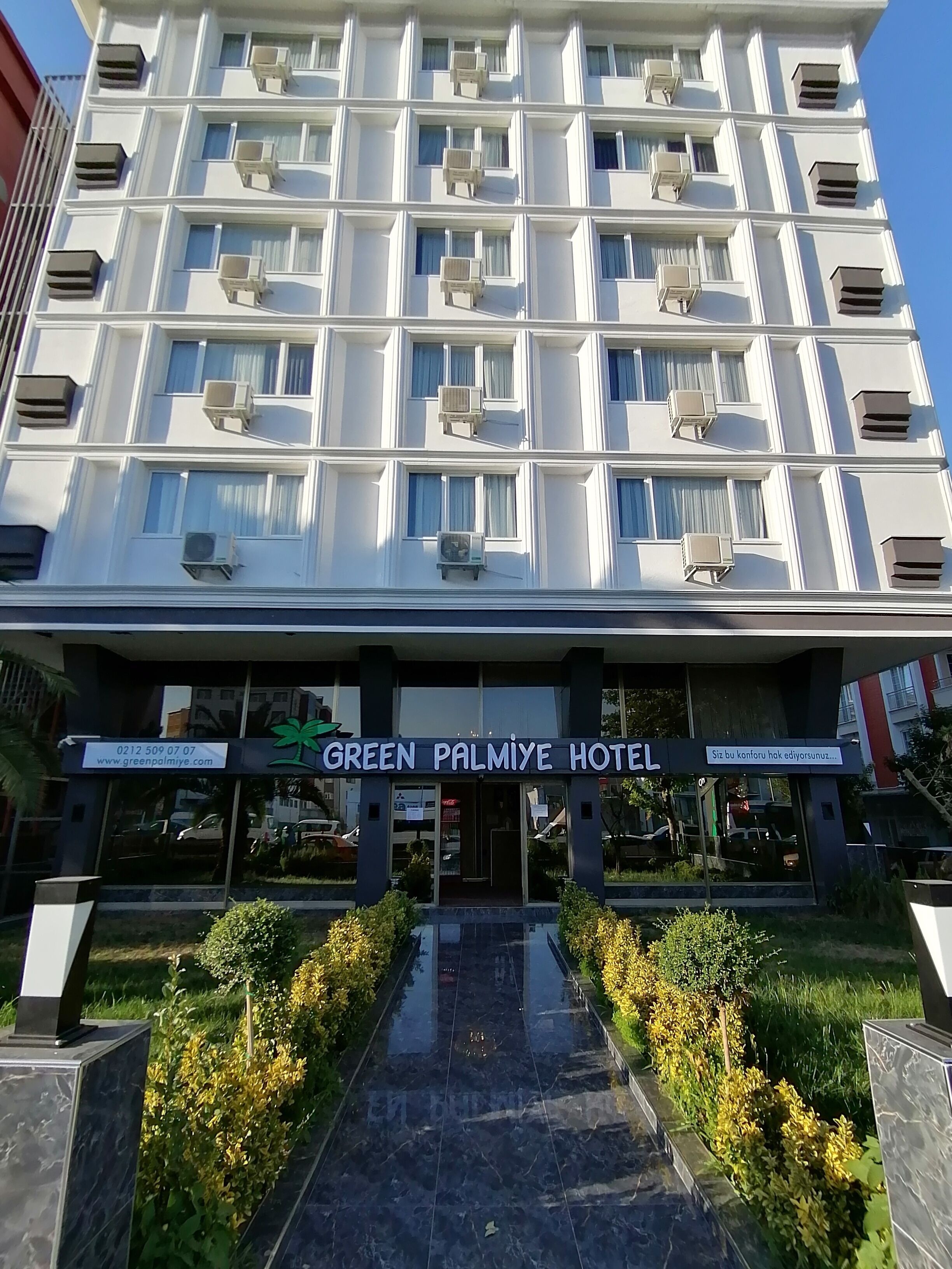 A multi-story hotel with air conditioning units on each floor, a well-maintained entrance with a glass door, and a signboard displaying the hotel name.