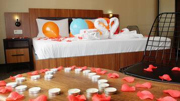 Habitación de hotel con una cama bien hecha, una mesita de noche, una silla y una mesa con velas y pétalos de rosa.