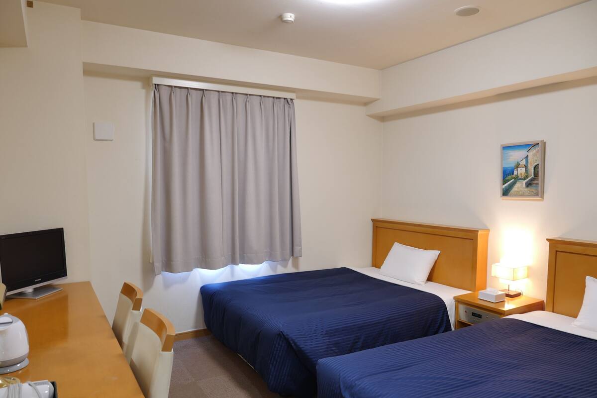 Una habitación de hotel con dos camas, un escritorio con computadora, una silla, un ventanal con cortinas y un cuadro en la pared.