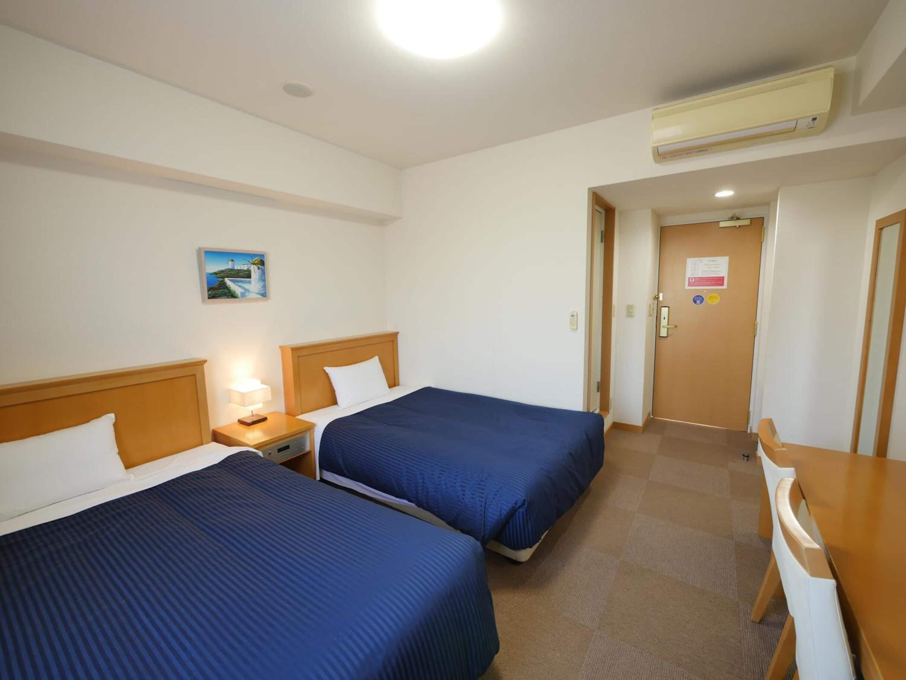 A hotel room with two beds, a desk, and a chair. There is a painting on the wall and an air conditioning unit above the bed.