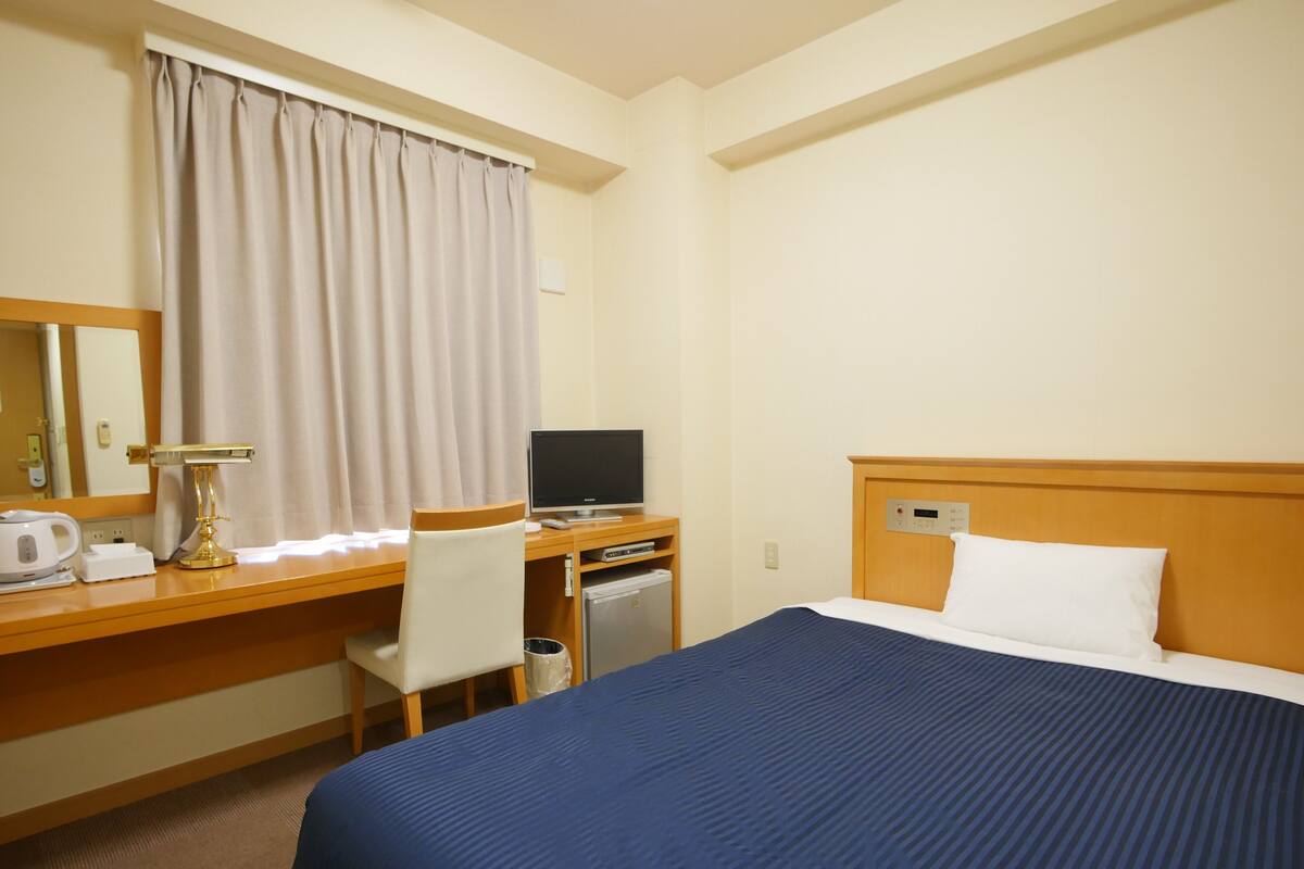 Habitación de hotel con cama, escritorio con computadora, silla, kitchenette pequeño y ventana con cortinas.