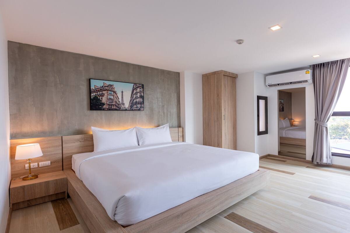 A modern hotel room with a large bed, a nightstand with a lamp, a painting of Paris, a wooden wardrobe, an air conditioning unit, and a view of the city through a large window.