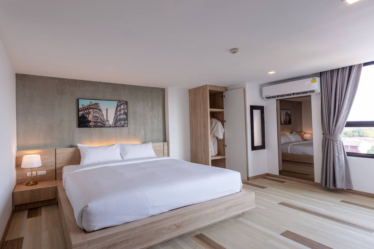 A modern hotel room with a large bed, a nightstand, a wall-mounted lamp, a wardrobe, and a window with curtains.