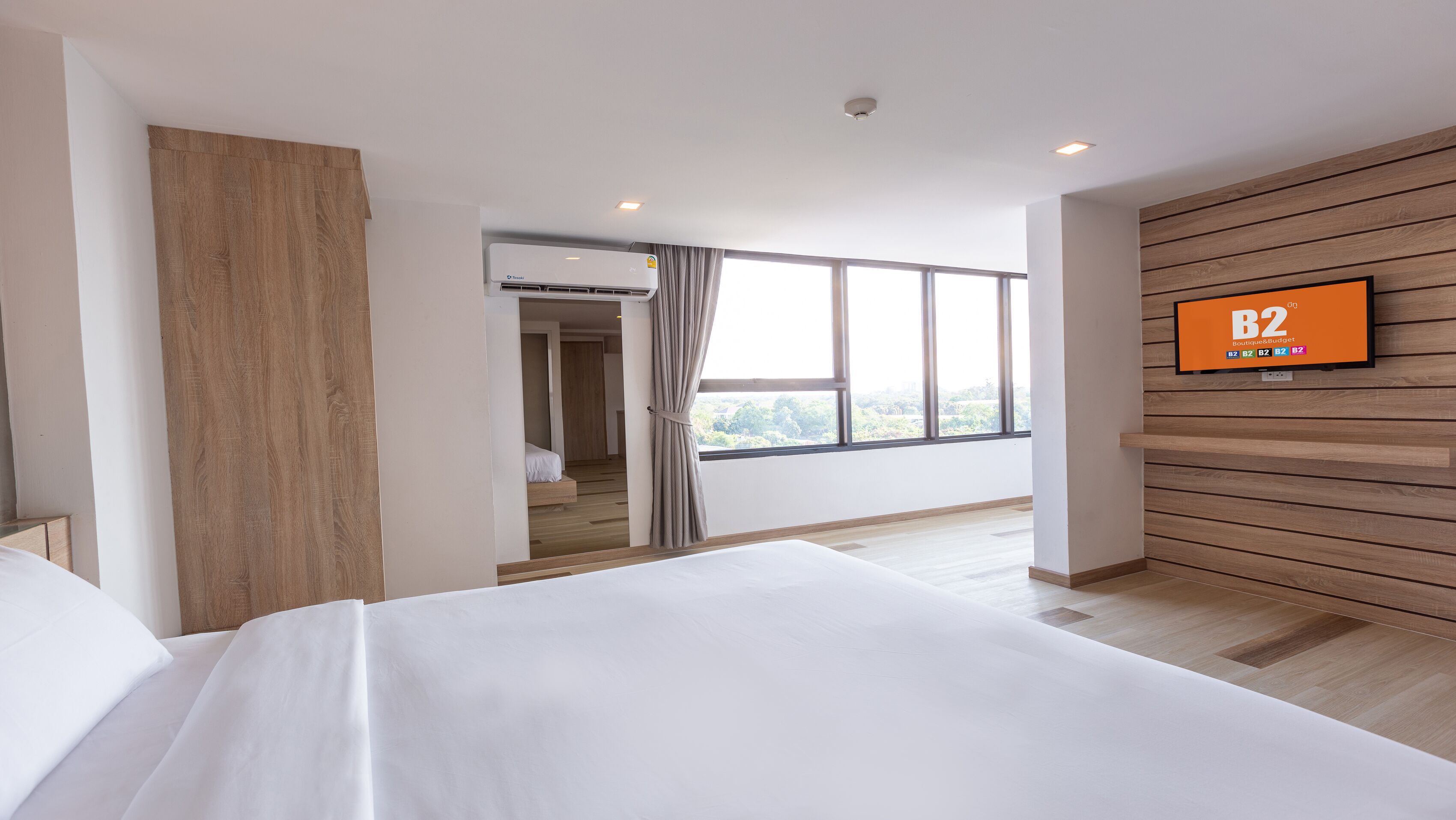 A modern hotel room with a large bed, a TV mounted on the wall, and a view of the city through large windows.