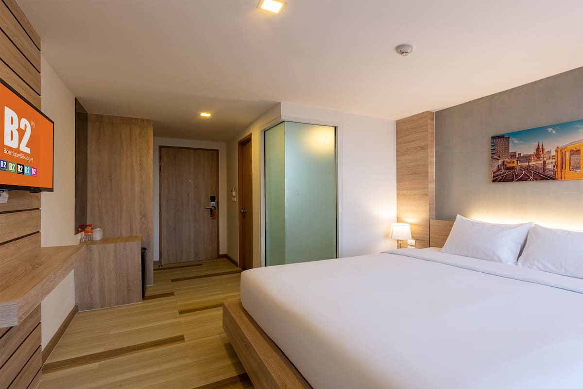 A hotel room with a large bed, a painting on the wall, a TV on the left, and a door leading to another room.