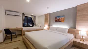 A hotel room with a large bed, a desk, a chair, a nightstand with a lamp, an air conditioning unit, and a cityscape painting on the wall.